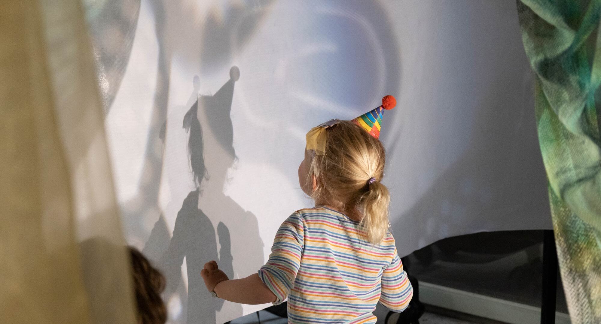 A young child seen from behind, wearing a pointy party hat and dancing in front of a wall with light patterns shining on it