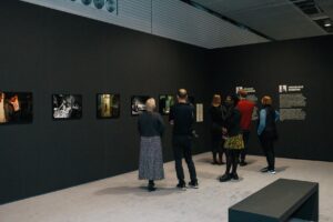 A small group of people looking at a photography exhibition. The photos are displayed on dramatic black walls.