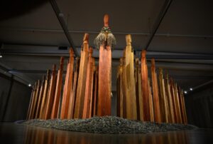 A sculpture installation. Many tall upright wooden columns with figurative faces carved on top, arranged in an arrow formation.