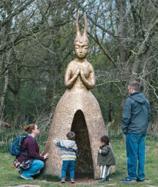 22-big-sunday-05.04.26-photo-by-kat-mager Two young children and two adults looking at a large outdoor sculpture made of bronze in the shape of a human figure with rabbit ears and hand held together in a prayer pose