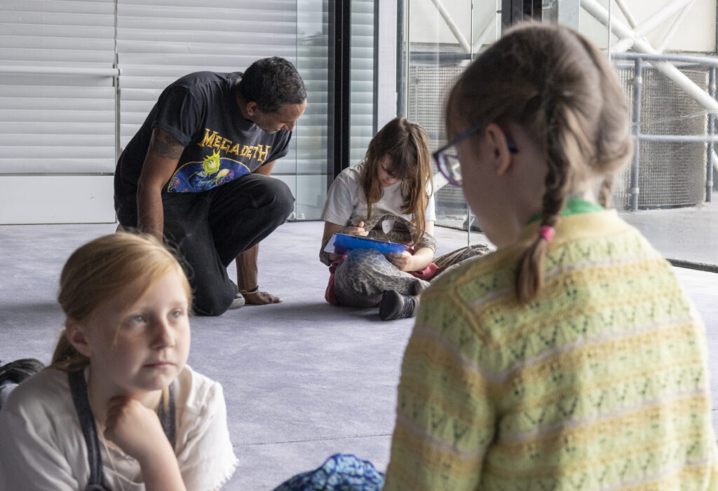 Older man helps young girl with her writing. Two other young girls sit in the foreground