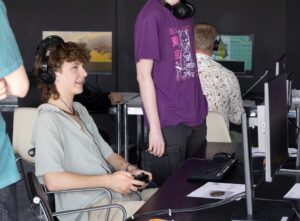A young person seen sideways playing a computer game on a desktop screen and wearign headphones. Other people are also playing games in the background.
