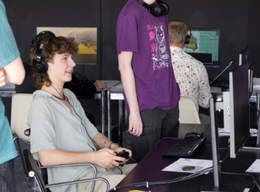 A young person seen sideways playing a computer game on a desktop screen and wearign headphones. Other people are also playing games in the background.