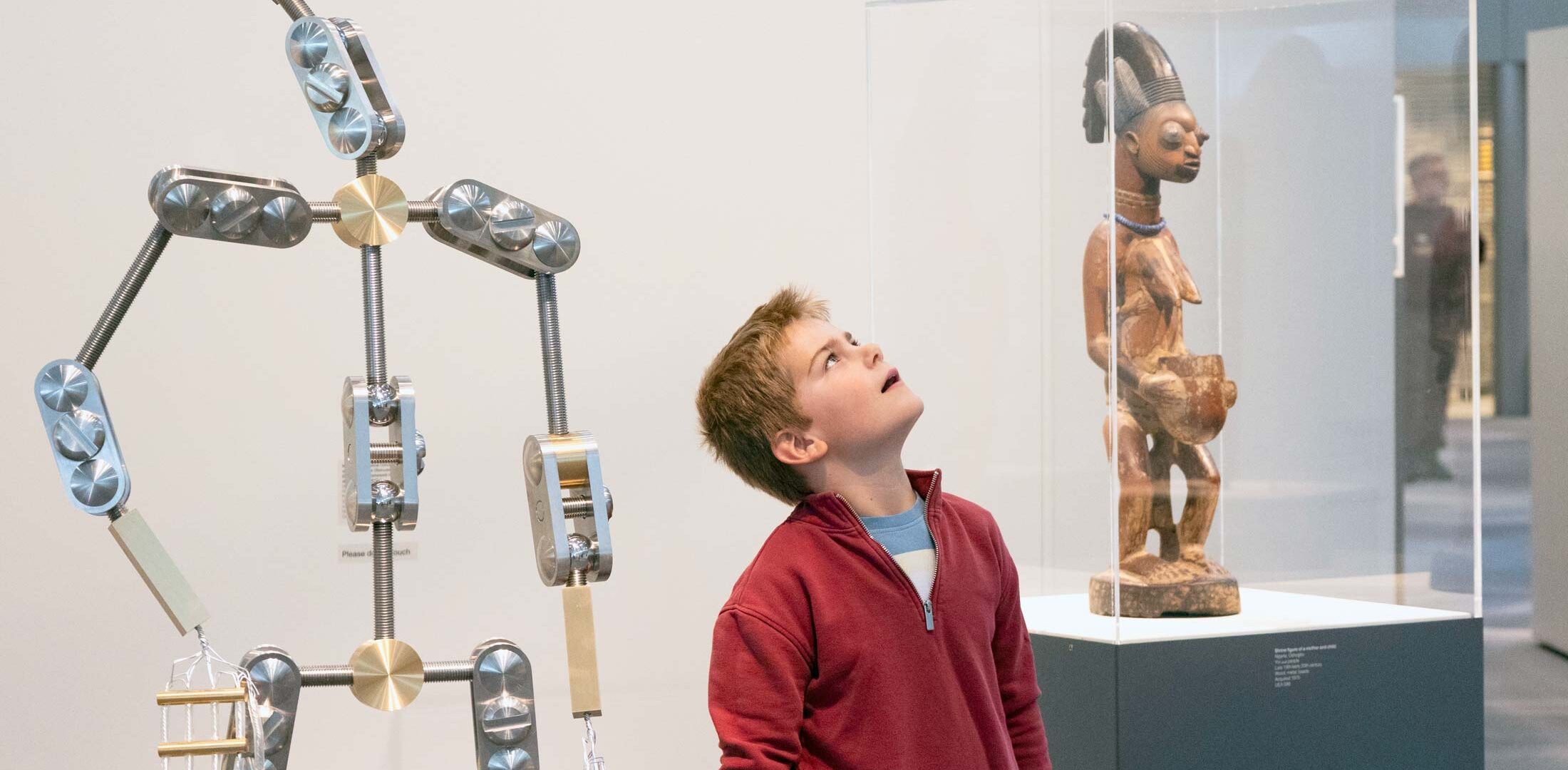 A young boy standing in an art gallery looking upwards next to a semi-figurative sculpture that is also looking upwards