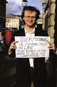 A young man wearing glasses and a black suit jacket with white shirt holding up a sign written in black pen on white paper that says EVERYTHING IS CONNECTED IN LIFE THE POINT IS TO KNOW IT AND TO UNDERSTAND IT