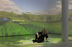 A person dressed in black posing on the floor in front of a large film projection in a gallery space. The film shows digital imagery of grass fields and woodland.