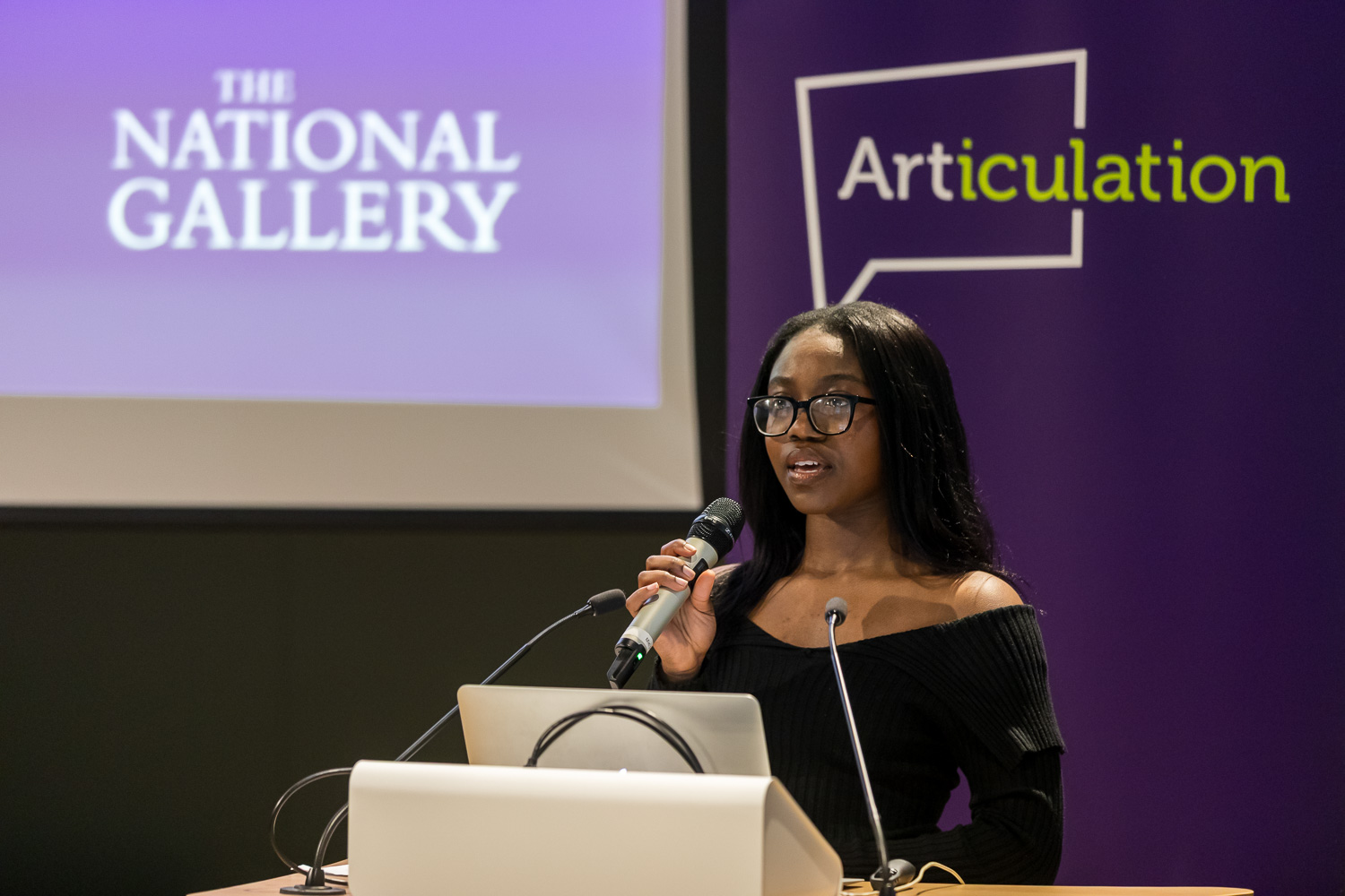 A dark skinned young lady standing ona podium with a mic in her hand and a purple roll up banner in the back with the Articulation logo on it. There is also the National Gallery logo on a projector in the background.