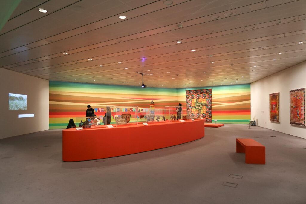 Installation view of an art exhibition with a large bright red plinth in the centre with objects on top and a brightly coloured wall mural behind with striped patterns.