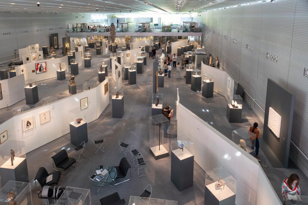 A view looking downwards onto a large exhibition area in an art museum, with objects on display and visitors looking at them.