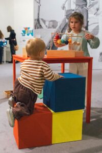 A young child seen from behind climbing on some coloured fabric blocks, looking at a table top, where another child is playing a game with some coloured knitted balls