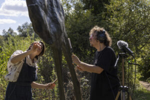 Two people tap a large bronze sculpture with small wooden drumsticks whilst recording the noise