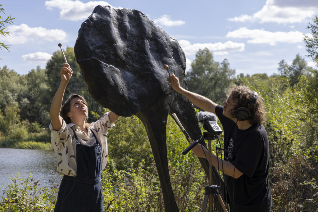 Two people tap a large bronze sculpture with small wooden drumsticks whilst recording the noise